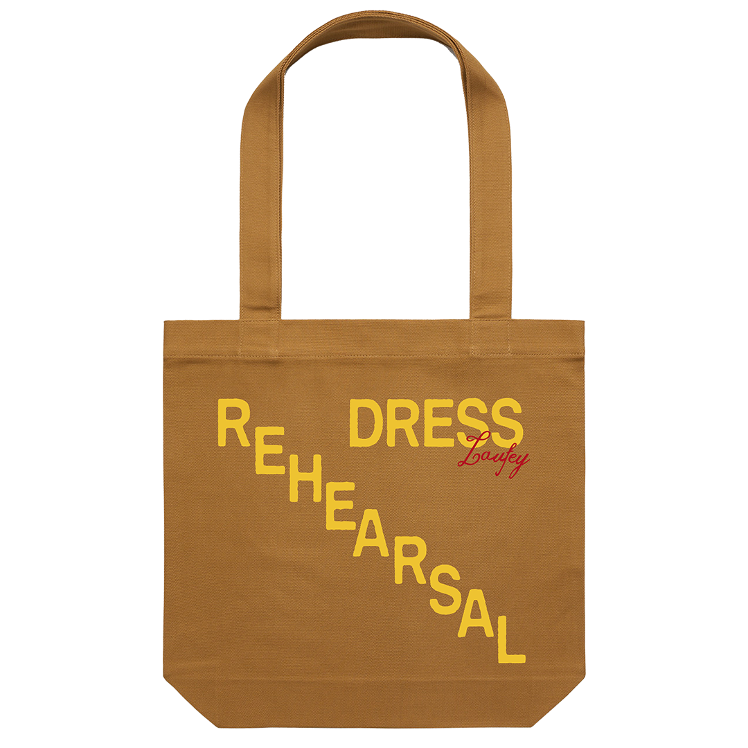 A camel-colored cotton tote bag with the words 'REHEARSAL' and 'DRESS' printed in a yellow gradient on the side.