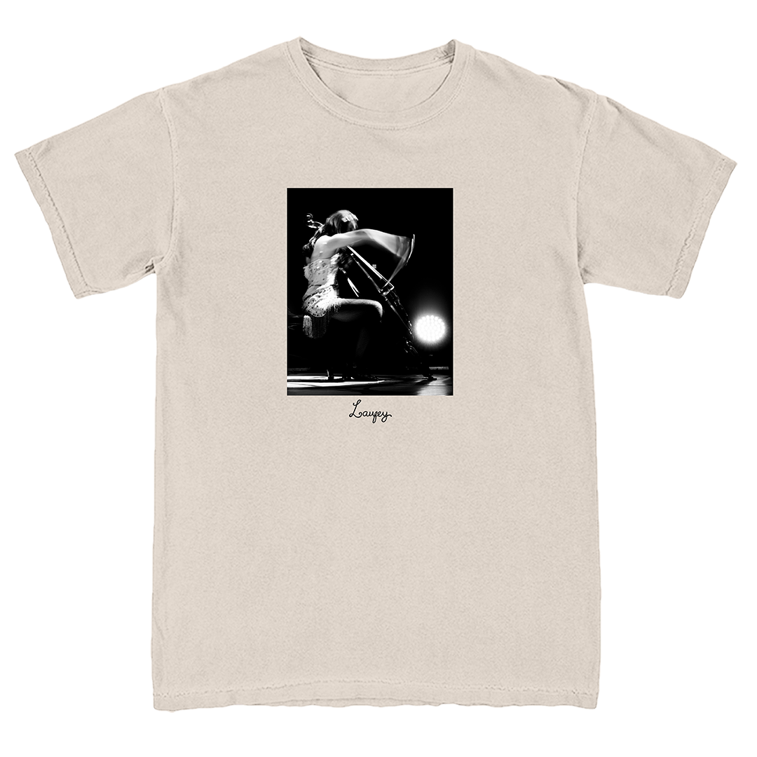 A light beige crewneck t-shirt featuring a black and white graphic print of a cellist on stage, with the name 'Loursy' printed in script below the image.