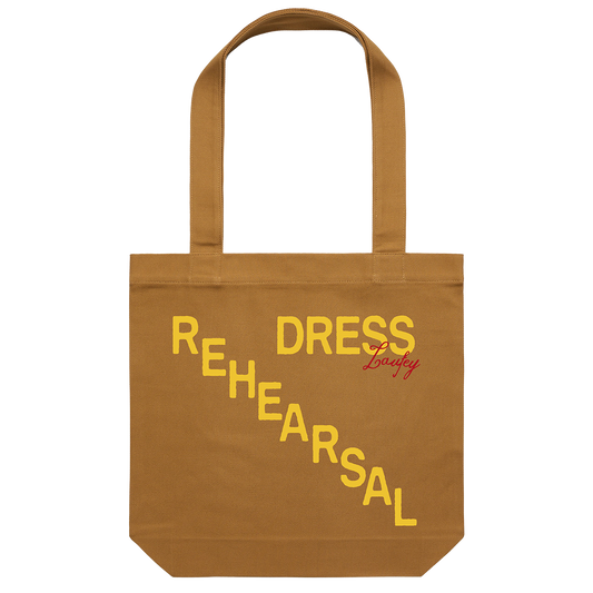 A camel-colored cotton tote bag with the words 'REHEARSAL' and 'DRESS' printed in a yellow gradient on the side.